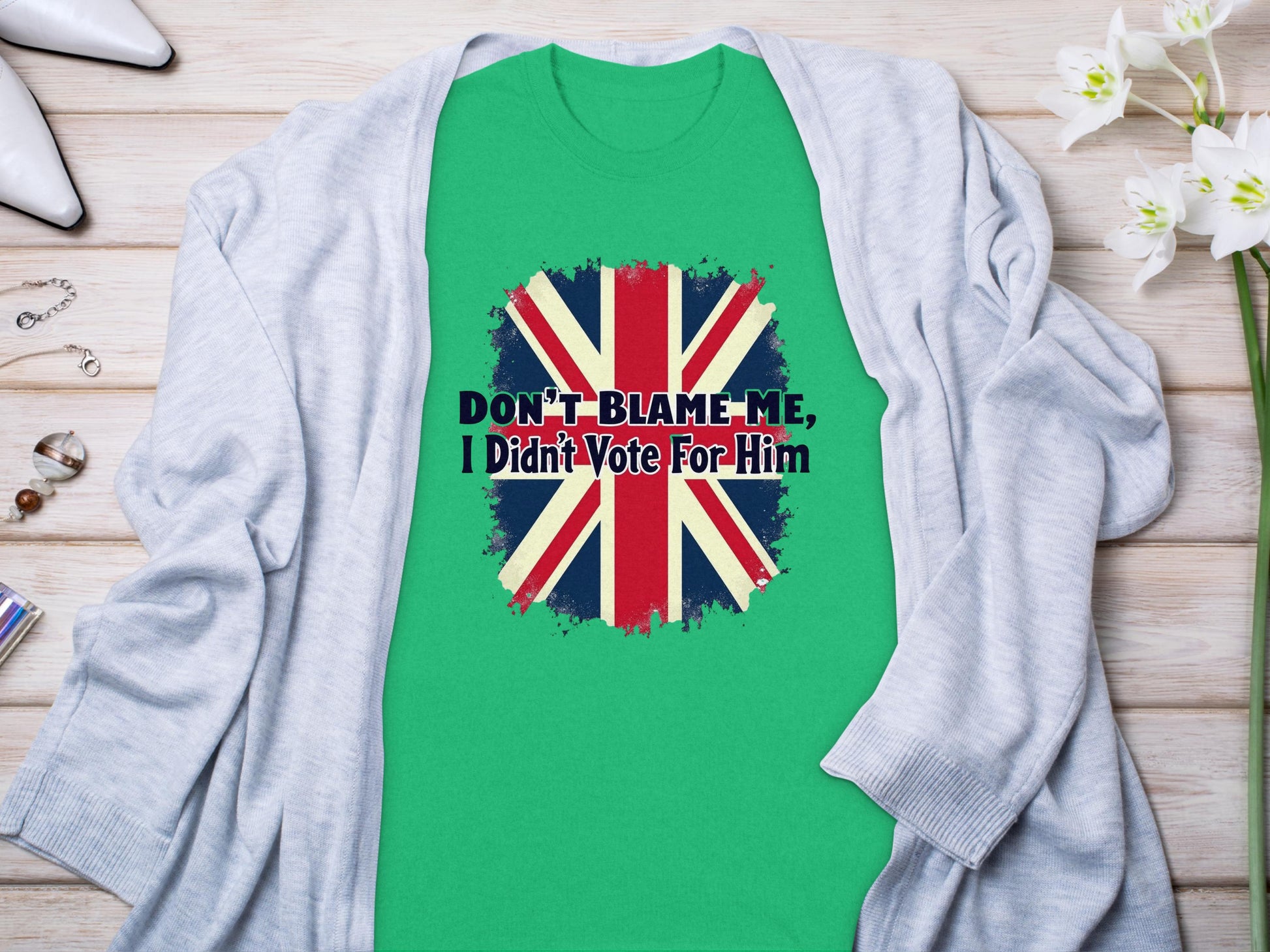 The classic fit Green T-shirt from Garment Graphics features the Union Jack and bold text: Dont Blame Me, I Didnt Vote for Him. It is displayed on a wooden surface with a light gray cardigan, white shoes, and flowers nearby.