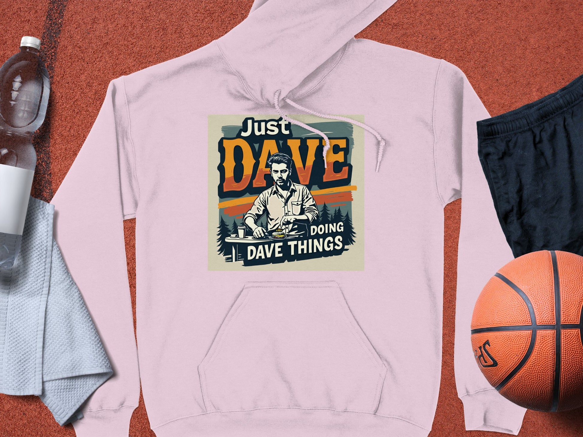 A light pink graphic hoodie from Garment Graphics features a man at a desk with Just Dave Doing Dave Things. Made of medium-heavy fabric, its displayed with a basketball, black shorts, water bottle, and gray towel.