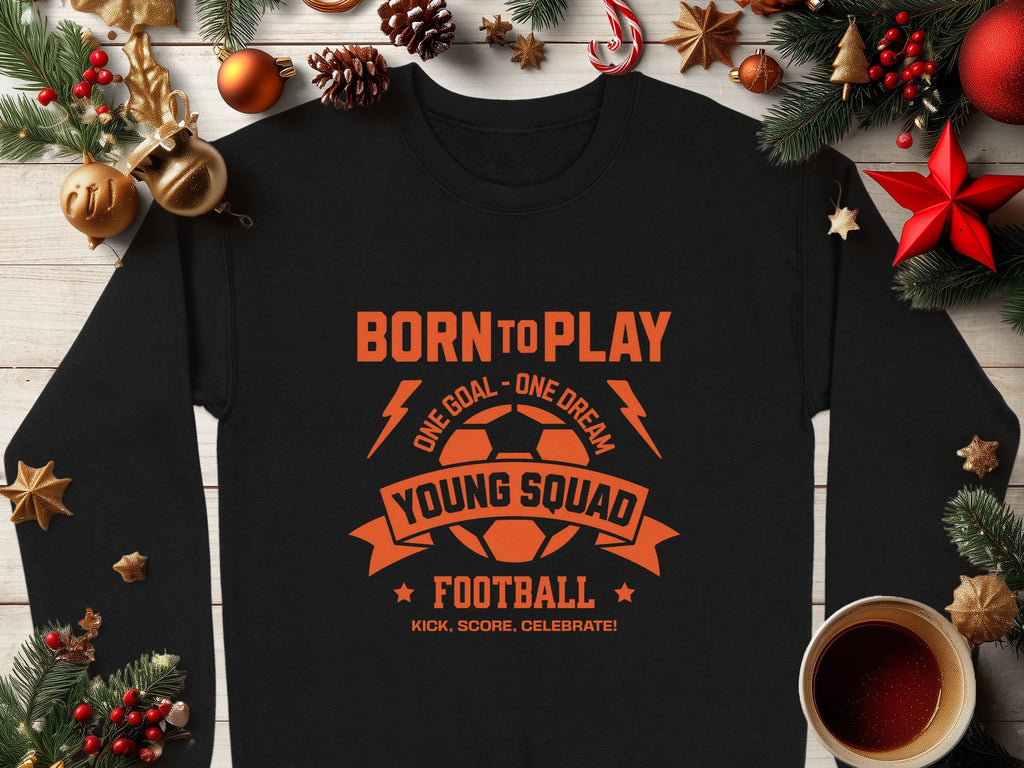 The black unisex sweatshirt by Garment Graphics showcases an orange text reading, BORN TO PLAY, ONE GOAL - ONE DREAM, YOUNG SQUAD, FOOTBALL, KICK, SCORE, CELEBRATE! alongside a soccer ball graphic. Its displayed with festive decor and a steaming cup on a wooden surface.