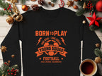 The black unisex sweatshirt by Garment Graphics showcases an orange text reading, BORN TO PLAY, ONE GOAL - ONE DREAM, YOUNG SQUAD, FOOTBALL, KICK, SCORE, CELEBRATE! alongside a soccer ball graphic. Its displayed with festive decor and a steaming cup on a wooden surface.