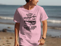 A person stands on a beach wearing a pink Garment Graphics T-shirt with a Classic London City Vintage Car Club design. The sandy beach, clear sky, and water elevate the retro automotive vibe of this classic fit attire.