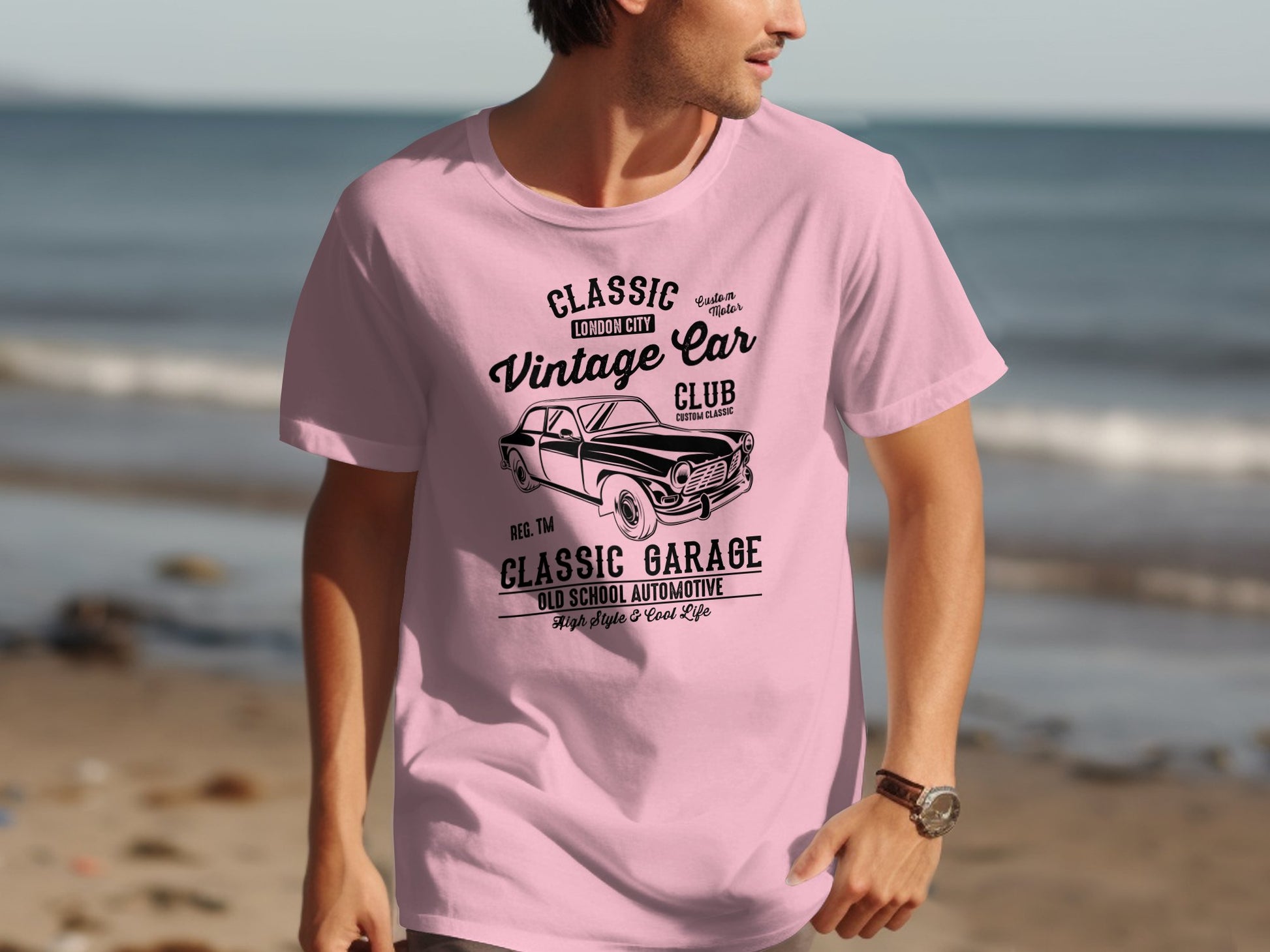 A person stands on a beach wearing a pink Garment Graphics T-shirt with a Classic London City Vintage Car Club design. The sandy beach, clear sky, and water elevate the retro automotive vibe of this classic fit attire.