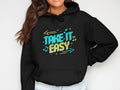 Someone is wearing a Garment Graphics classic fit black hoodie, showcasing colorful text and graphics with the phrase Take It Easy in bright blue and yellow, enhanced by musical notes and decorative arrows. They are smiling, showing just the lower half of their face.