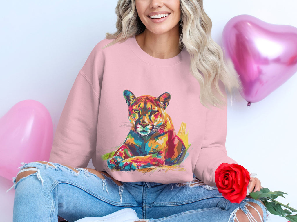 A person smiles, seated on the floor, holding a red rose. They wear Garment Graphics pink sweatshirt with a vibrant big cat design and ripped jeans. Two pink heart-shaped balloons float in the background, enhancing the cheerful atmosphere.