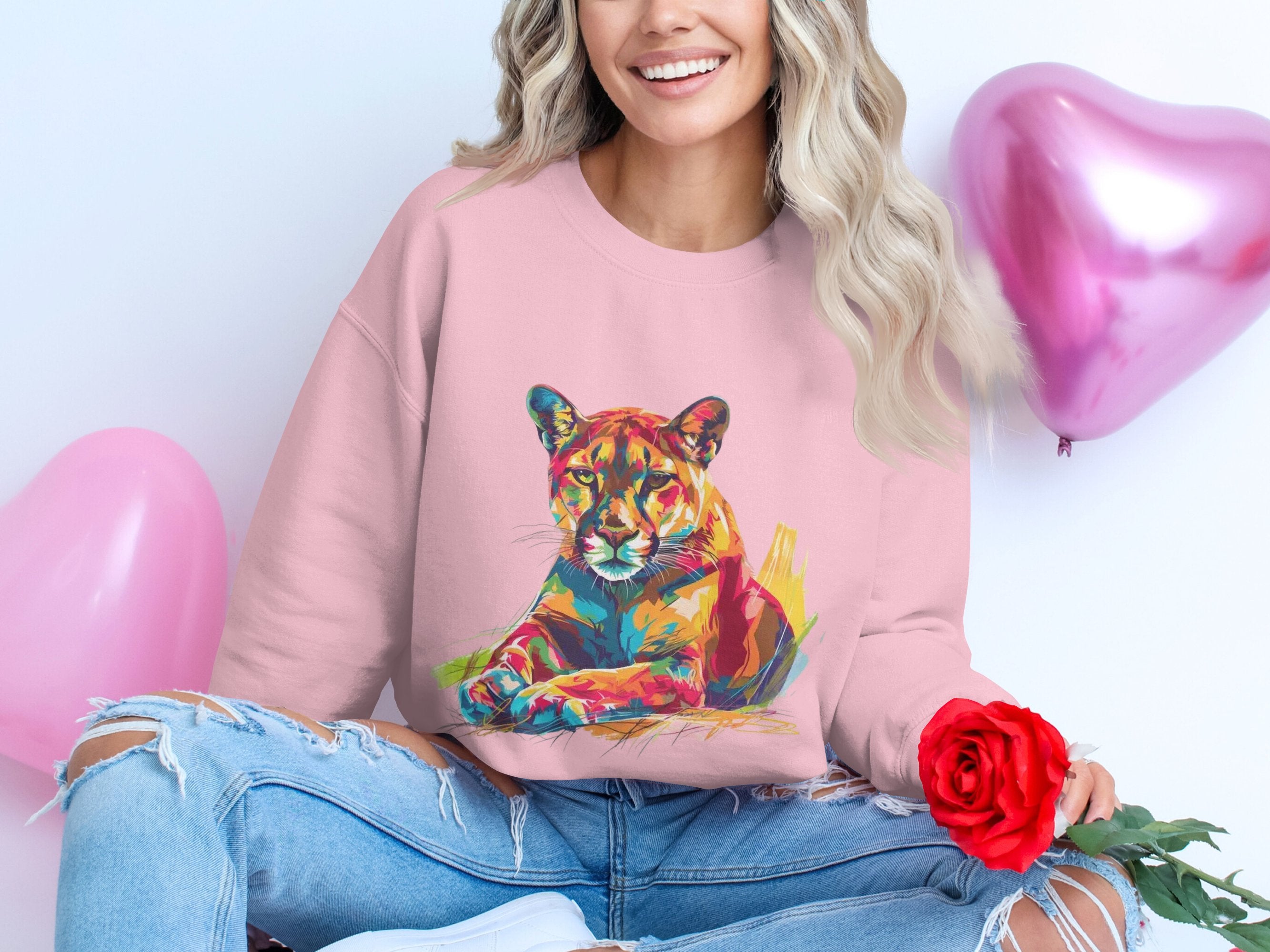 A person smiles, seated on the floor, holding a red rose. They wear Garment Graphics pink sweatshirt with a vibrant big cat design and ripped jeans. Two pink heart-shaped balloons float in the background, enhancing the cheerful atmosphere.