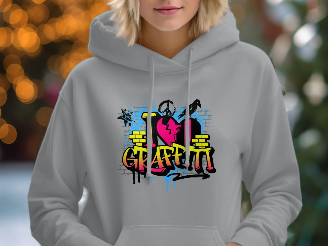 The Garment Graphics hoodie with I Love Graffiti in blue, yellow, and pink splashes lies on a wooden surface. Ideal for any casual wardrobe, its surrounded by a hat, sunglasses, shoes, a notebook, and a camera—capturing perfect urban street art vibes.