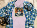 A Garment Graphics lavender hoodie with Just Dave Doing Dave Things text and a man graphic, made from medium-heavy fabric, is displayed on wood with a blue plaid shirt, headphones, camera, and water bottle.