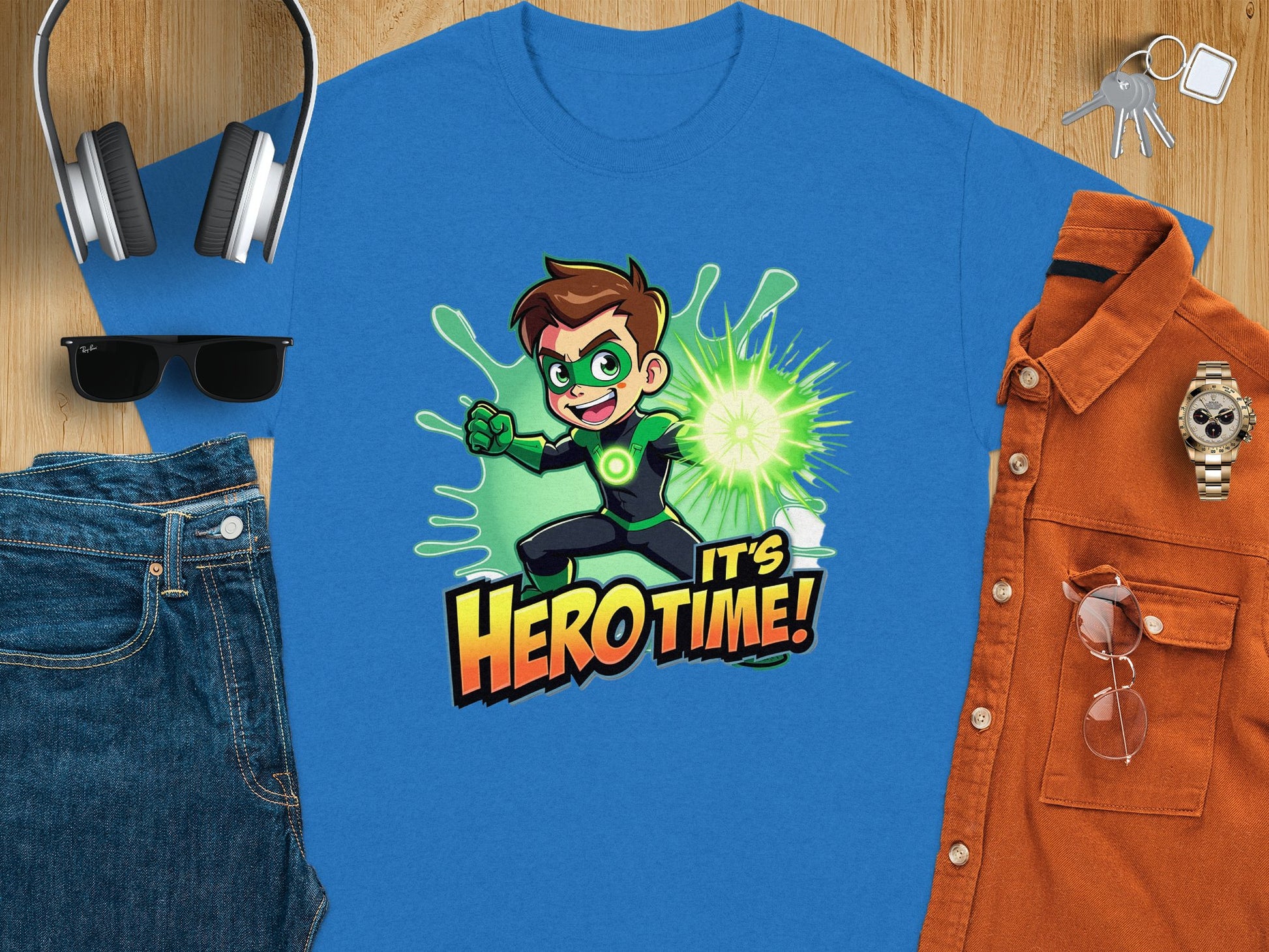 Garment Graphics offers a vibrant blue t-shirt with a superhero print—featuring brown hair, green mask and gloves, and the phrase Its Hero Time! by a neon green explosion. Nearby are headphones, sunglasses, blue jeans, an orange shirt, and a wristwatch on a wooden surface. Perfect for family fun!.
