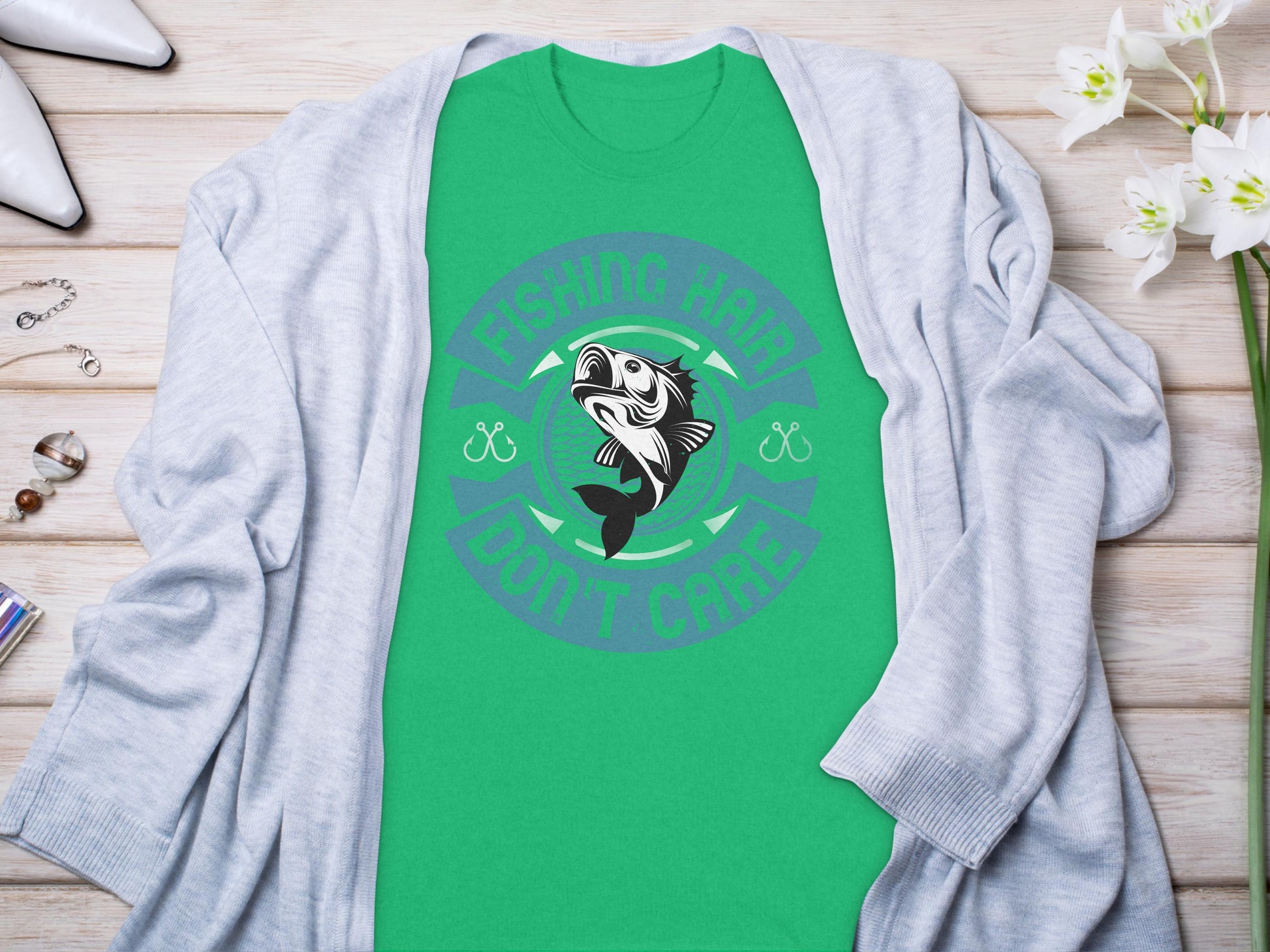 A green Garment Graphics tee features a unique fish design with the text Fishing Hard, Dont Care. It lies on a light wooden surface, accompanied by a gray cardigan, white flowers, jewelry, and sunglasses.