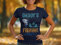 A person wears a Garment Graphics navy fishing T-shirt with Daddys Fishing Buddy text, featuring a fish and boater against autumn trees. Hands on hips, it captures the essence of leisure and style for fishing enthusiasts.