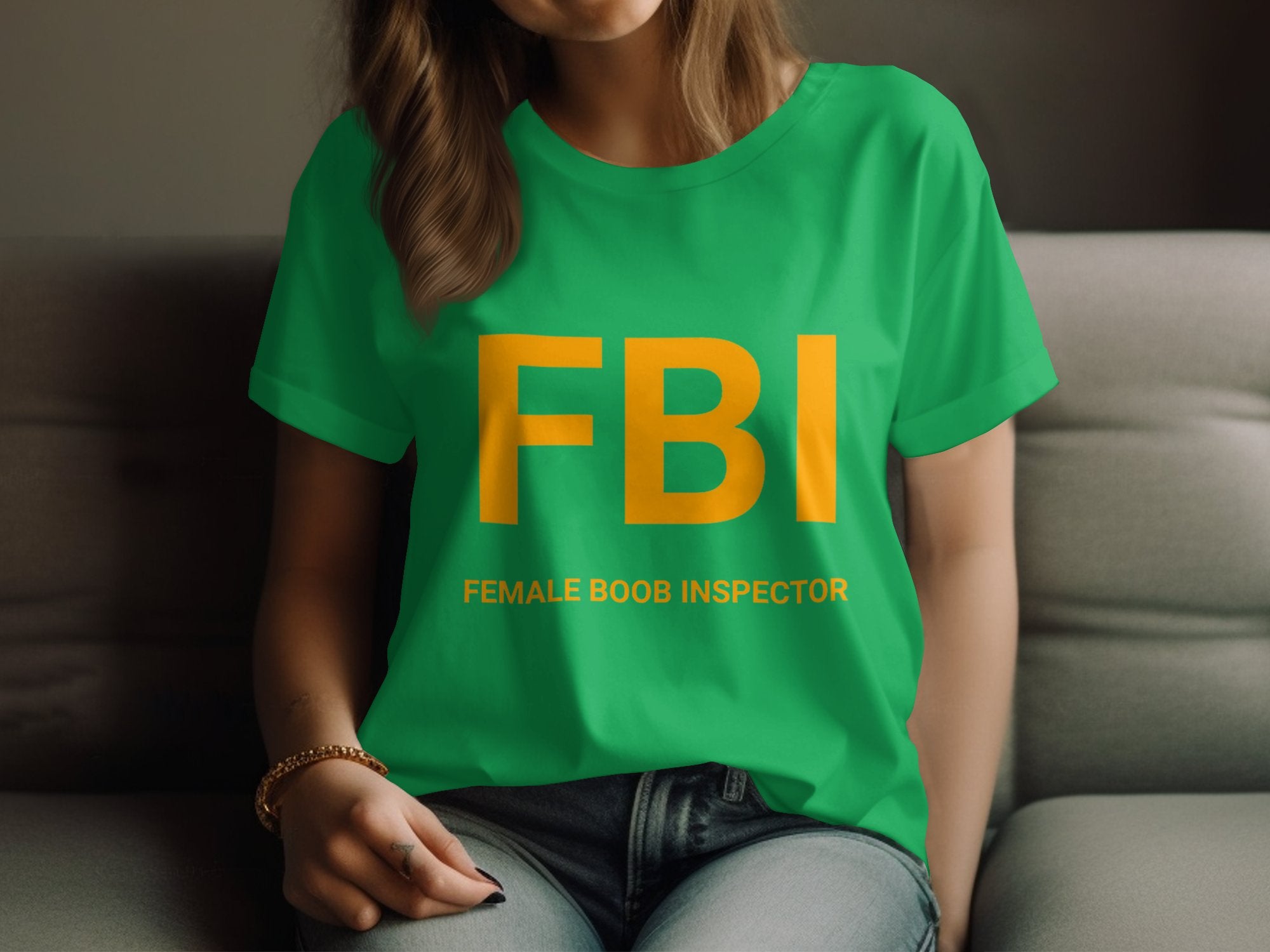 A person on a sofa wears a Garment Graphics green T-shirt with large yellow FBI letters above Female Boob Inspector. The womens fit complements their long hair as they hold their knee with one hand, making it a classic gag gift.