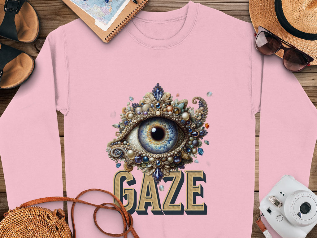A pink unisex graphic sweatshirt by Garment Graphics features an ornate jeweled eye design above GAZE in bold. Its styled with a sun hat, sunglasses, a notebook, a map, a camera, and sandals on a wooden surface.