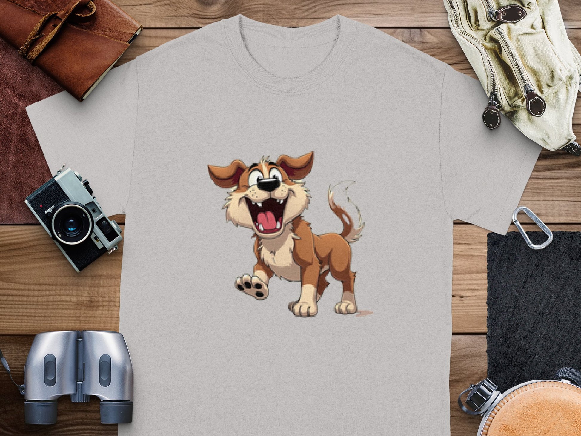 A light gray T-shirt by Garment Graphics lies on a wooden surface with a cartoon of a happy brown dog, ideal for dog lovers. Around the shirt are a wallet, camera, gloves, binoculars, and other items.