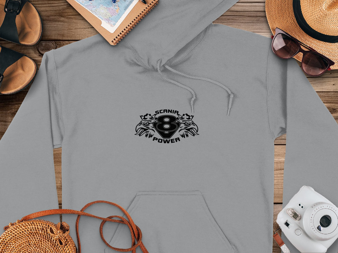 A white hoodie by Garment Graphics features a bold SCANIA V8 POWER graphic on the front and a classic fit. Its styled on a wooden surface with a notebook, straw hat, glasses, instant camera, and woven bag—perfect for Scania Power Eight Hoodies fans.