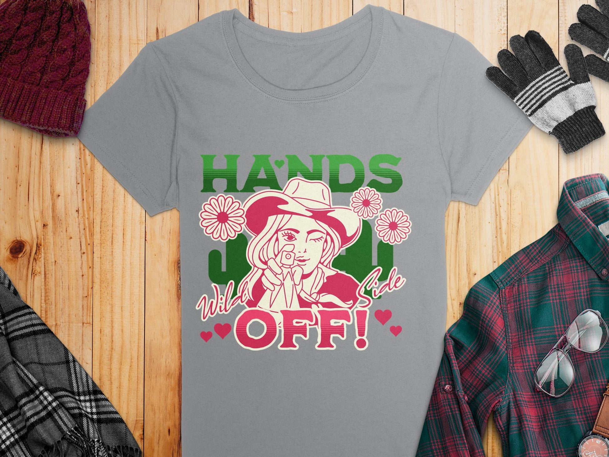 A stylish gray T-shirt from Garment Graphics features a bold design of a woman with a hat and sunglasses, accented by daisies and the text Hands Off! Wild Side. Its surrounded by a plaid scarf, knit hat, gloves, and plaid shirt on a wooden surface.