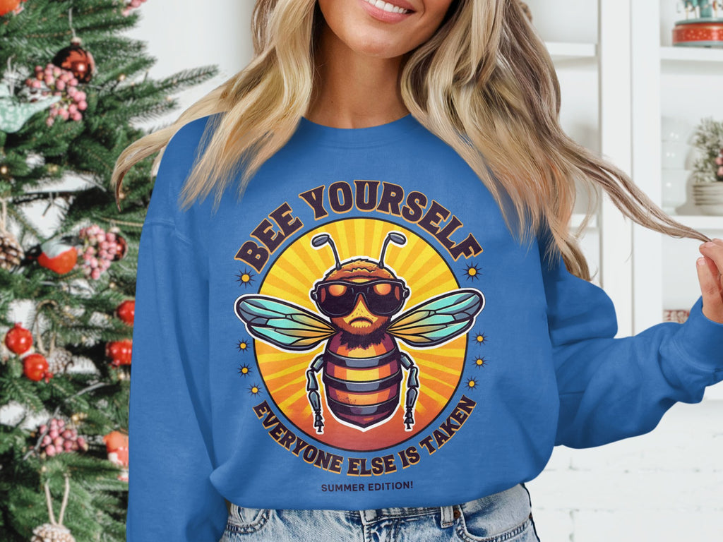 The person is wearing a Garment Graphics Summer Edition Sweatshirt in blue, unisex sizing, featuring a bee with sunglasses and the phrase Bee Yourself, Everyone Else is Taken in a circular design. A decorated Christmas tree adds festive flair to the left background.