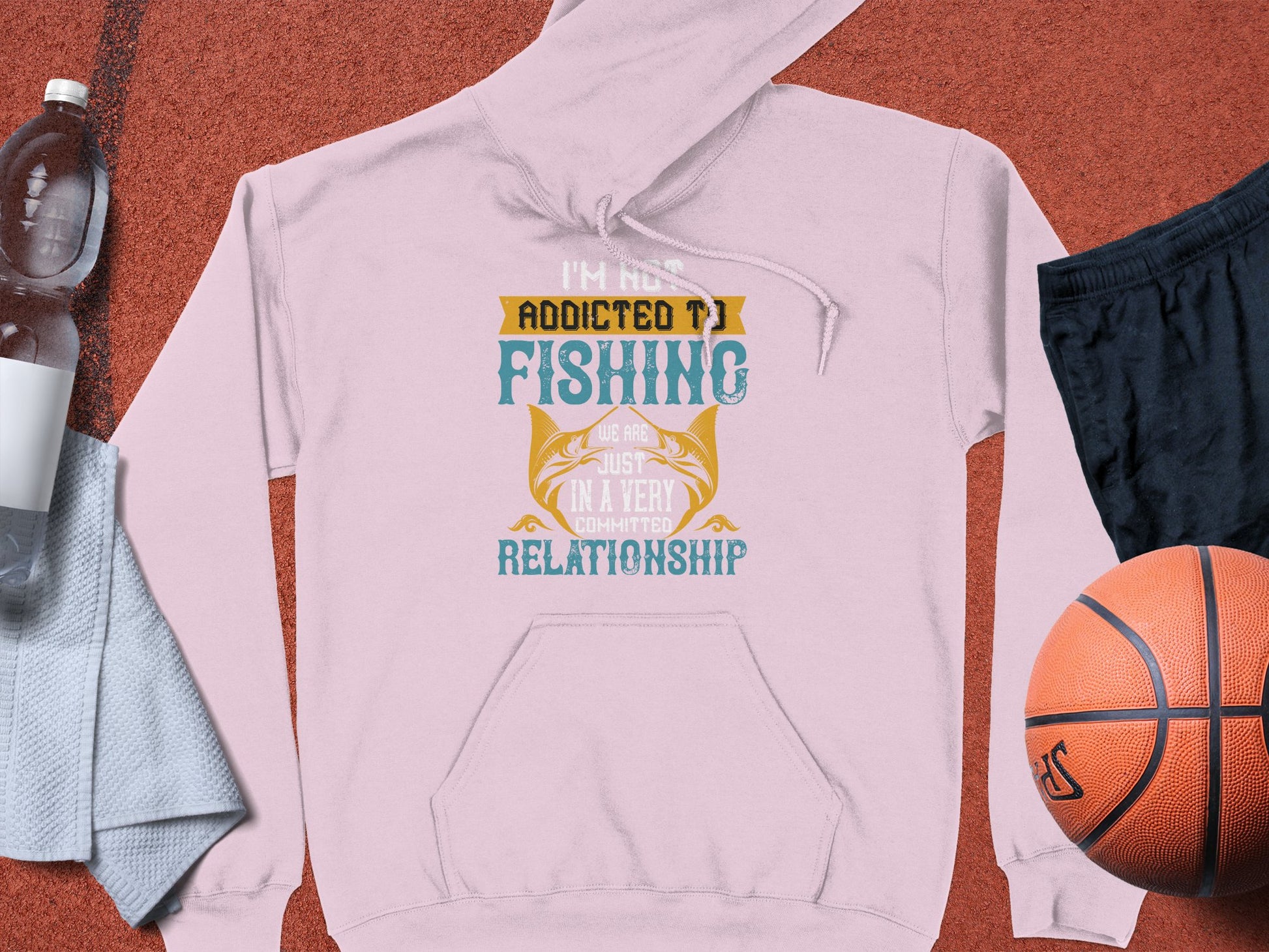 A classic fit light pink fishing hoodie by Garment Graphics lies on the sports court, showcasing the text Im not addicted to fishing, were just in a very committed relationship. Nearby are a basketball, black shorts, a water bottle, and a blue towel.