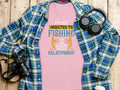 The Garment Graphics pink fishing t-shirt, featuring the slogan Im not addicted to fishing, were just in a very committed relationship, is paired with a plaid shirt and accessorized with a camera and headphones on a wooden surface, perfect for avid anglers.