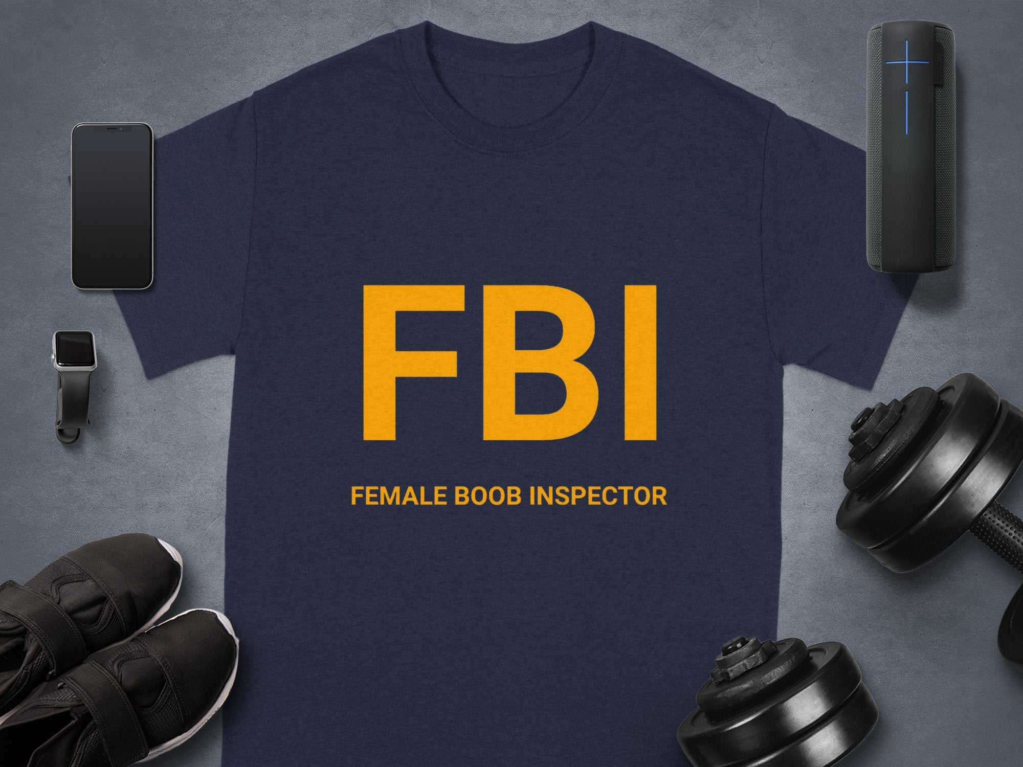 A navy blue Female Boob Inspector T-shirt by Garment Graphics flaunts bold yellow FBI letters and pairs hilariously with a smartphone, fitness tracker, black shoes, dumbbells, and a portable speaker—ideal for fans of funny designs.