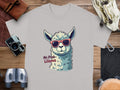 Garment Graphics offers a family-sized Llama T-Shirt featuring a cartoon llama in sunglasses and No Prob-Llama text. Displayed on wood, its surrounded by adventure gear such as cameras, binoculars, a bag, and a leather pouch.