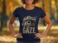 A woman in a navy blue Garment Graphics Gaze T-shirt, showcasing an ornate eye design with GAZE below, stands against a backdrop of vibrant autumn foliage.