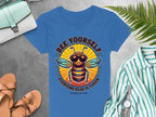 Garment Graphics womens blue t-shirt showcases a bee in sunglasses and reads Bee Yourself, Everyone Else is Taken, Summer Edition. Pair it with a striped shirt, sandals, watch, fern leaves, and bags on a gray surface for the perfect look.