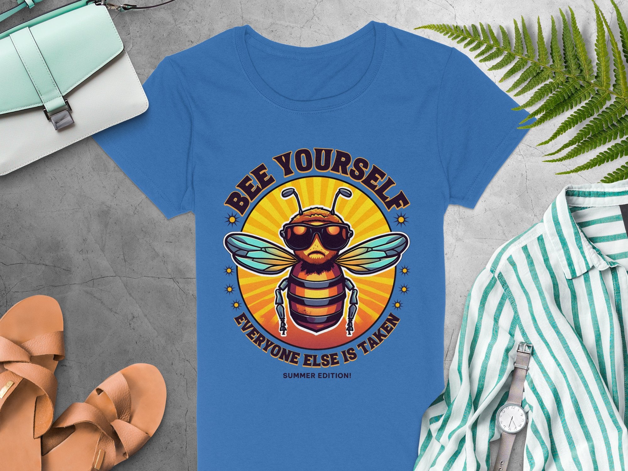 Garment Graphics womens blue t-shirt showcases a bee in sunglasses and reads Bee Yourself, Everyone Else is Taken, Summer Edition. Pair it with a striped shirt, sandals, watch, fern leaves, and bags on a gray surface for the perfect look.