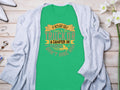 A Garment Graphics green t-shirt featuring the witty design If hes got golf clubs in his truck or a camper in his driveway... dont hire him is displayed on a wooden surface, accented by a gray cardigan, flowers, and accessories. Ideal for both camping and golf fans.