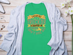 A Garment Graphics green t-shirt featuring the witty design If hes got golf clubs in his truck or a camper in his driveway... dont hire him is displayed on a wooden surface, accented by a gray cardigan, flowers, and accessories. Ideal for both camping and golf fans.