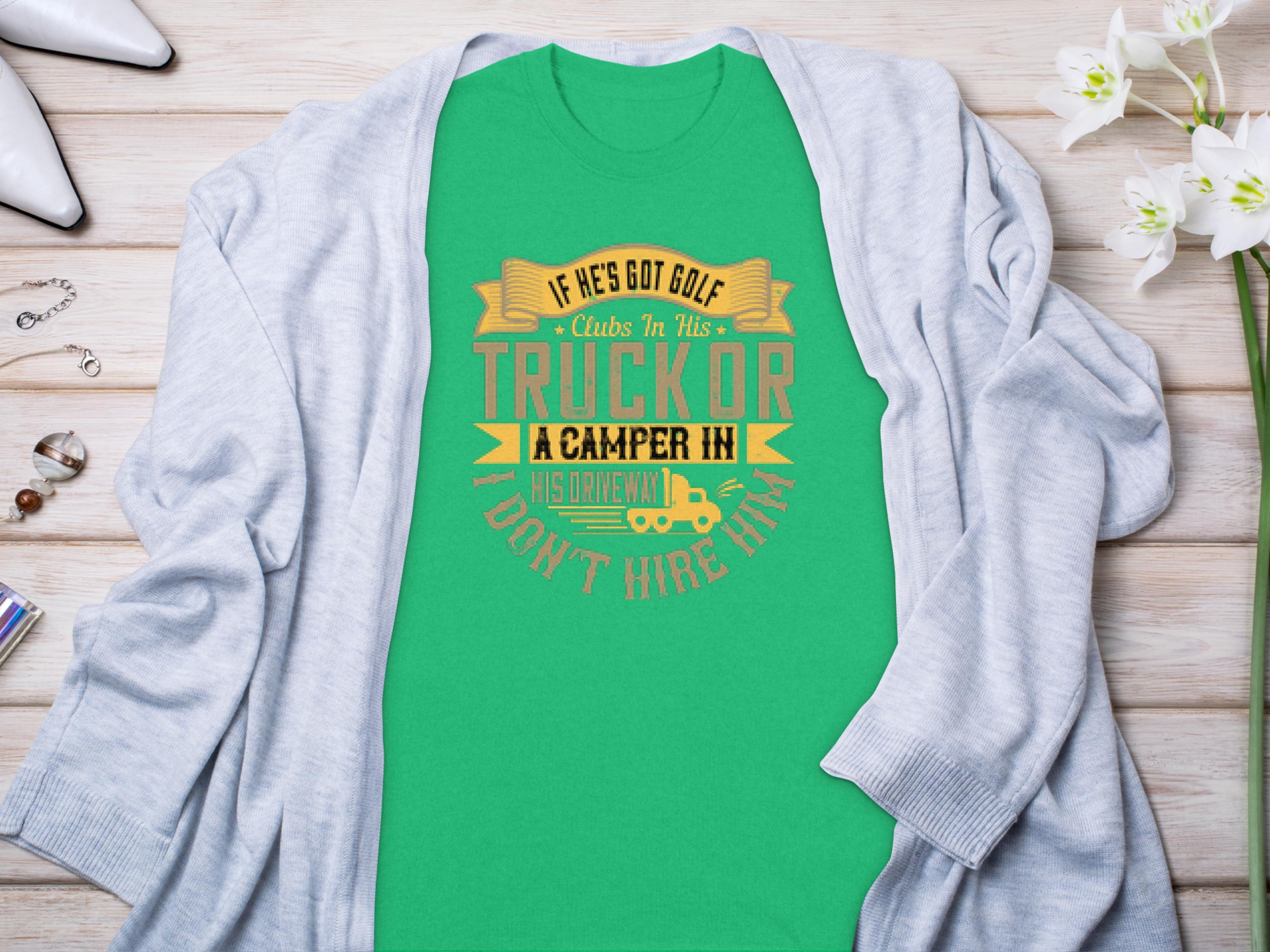 A Garment Graphics green t-shirt featuring the witty design If hes got golf clubs in his truck or a camper in his driveway... dont hire him is displayed on a wooden surface, accented by a gray cardigan, flowers, and accessories. Ideal for both camping and golf fans.
