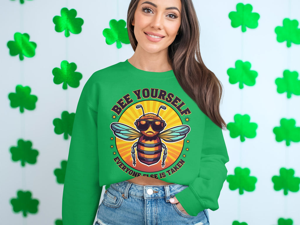 A person wears a green sweatshirt from Garment Graphics Summer Edition, featuring a bee illustration and the text Bee Yourself, Everyone Else is Taken, against a wall with four-leaf clovers. With unisex sizing, its ideal for anyone celebrating their unique buzz.