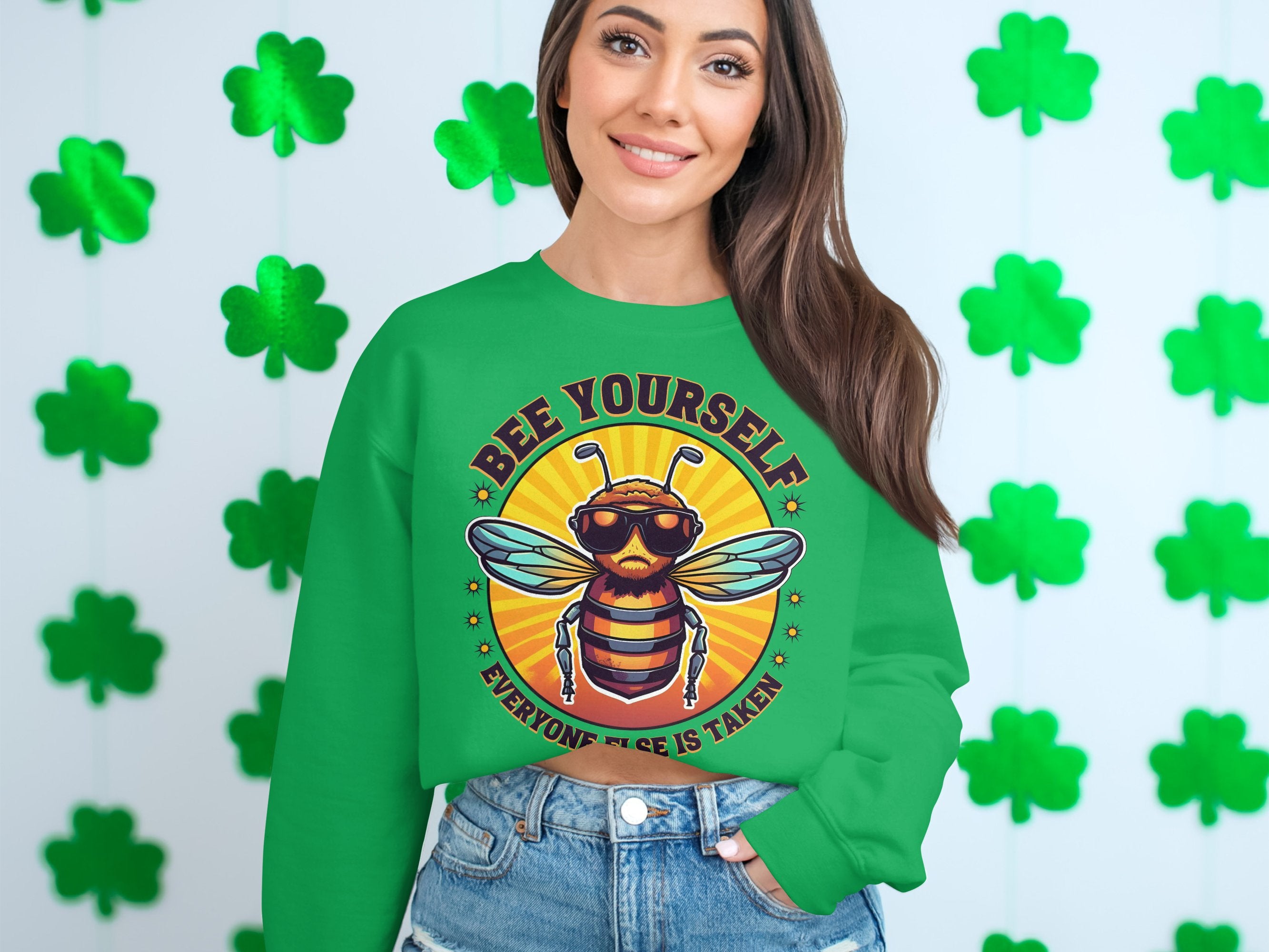 A person wears a green sweatshirt from Garment Graphics Summer Edition, featuring a bee illustration and the text Bee Yourself, Everyone Else is Taken, against a wall with four-leaf clovers. With unisex sizing, its ideal for anyone celebrating their unique buzz.