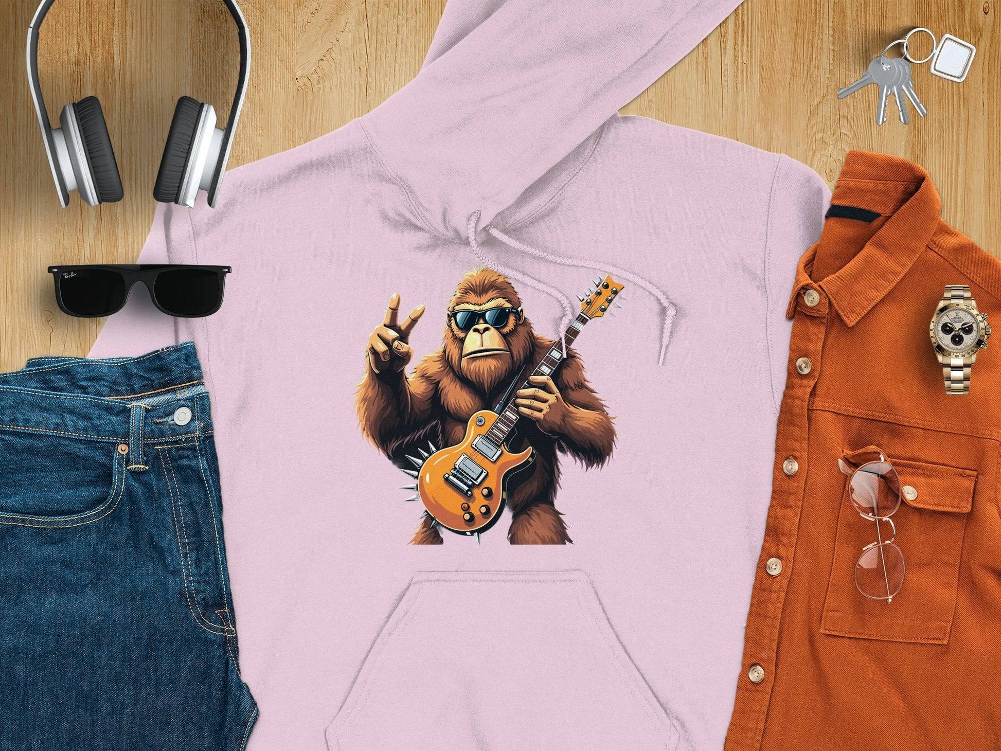 Garment Graphics flat lay: a gorilla hoodie with gorilla playing electric guitar surrounded by jeans, headphones, watch, keys, brown shirt with glasses, and sunglasses on wooden surface—ideal for music lovers wanting to rock their style!.