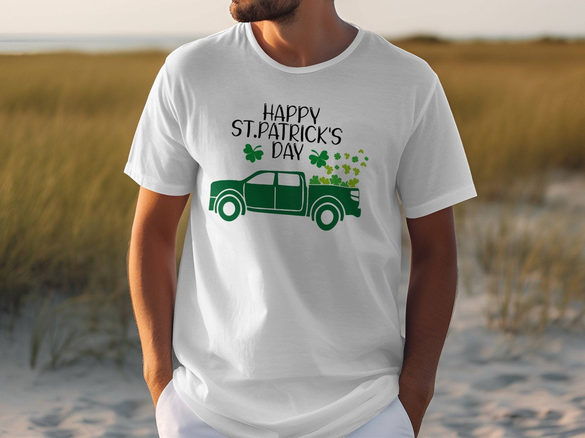 Someone sports a Garment Graphics white T-shirt featuring a green pickup truck filled with shamrocks. The shirt reads Happy St. Patricks Day, celebrating Irish pride, set against a grassy field backdrop.