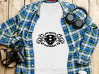 A white sweatshirt featuring Garment Graphics Scanir 8 Power logo and tribal designs is on a wooden surface, surrounded by a blue and green plaid shirt, black headphones, a camera, a cup, and a pen.
