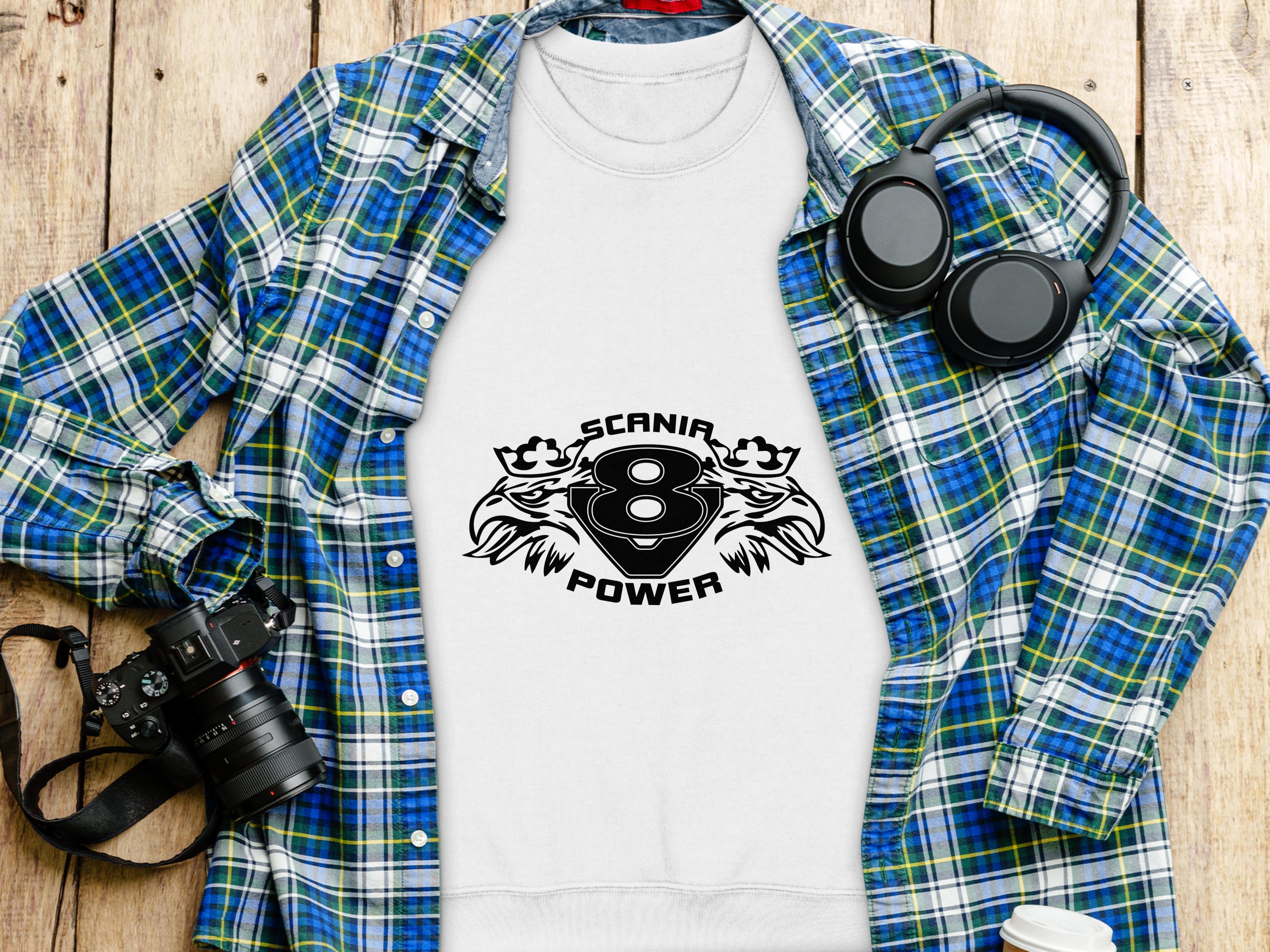 A white sweatshirt featuring Garment Graphics Scanir 8 Power logo and tribal designs is on a wooden surface, surrounded by a blue and green plaid shirt, black headphones, a camera, a cup, and a pen.