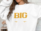 The individual wears a standout Garment Graphics sweatshirt displaying BIG in bold yellow letters, accompanied by two stylized eye graphics. Their long brown hair partially hides their face, enhancing the intrigue of this striking statement piece.