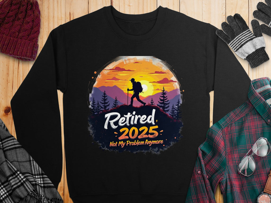 Garment Graphics offers a black retirement sweatshirt featuring a hiker silhouette against a sunset and trees. The design includes Retired 2025 Not My Problem Anymore. It pairs well with unisex-sized plaid shirts, scarves, hats, and gloves on a wooden surface.