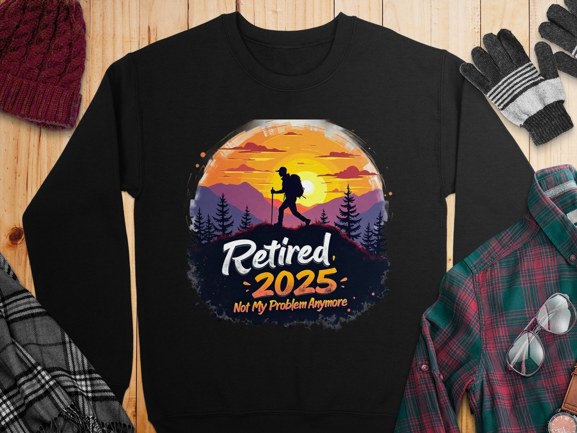 Garment Graphics offers a black retirement sweatshirt featuring a hiker silhouette against a sunset and trees. The design includes Retired 2025 Not My Problem Anymore. It pairs well with unisex-sized plaid shirts, scarves, hats, and gloves on a wooden surface.