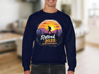 Someone wears a Garment Graphics unisex sweatshirt in navy blue featuring a hiker design with a sunset, trees, and the text Retired 2025 - Not My Problem Anymore. The room interior is softly focused.