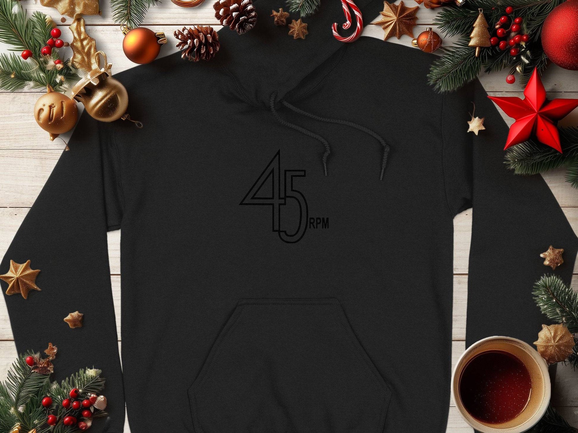 A black 45 RPM Retro Music Hoodie by Garment Graphics rests on a wooden surface, surrounded by festive pine cones, red and gold ornaments, star-shaped cookies, and a cup of black coffee—ideal for vinyl record enthusiasts.