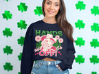 A woman stands by a wall of green shamrocks, sporting a black crop top from Garment Graphics Hands Off Wild Side collection. The sweatshirt bears HANDS OFF! with a bubble-blowing design and floral accents, combining boldness with a relaxed fit.