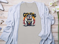 A gray Garment Graphics t-shirt with a crowned bulldog design and CITY KING text is on a light wood surface. Its paired with a light gray cardigan, white high heels, silver bracelet, and white flowers, highlighting the unique style of City King T-Shirts.