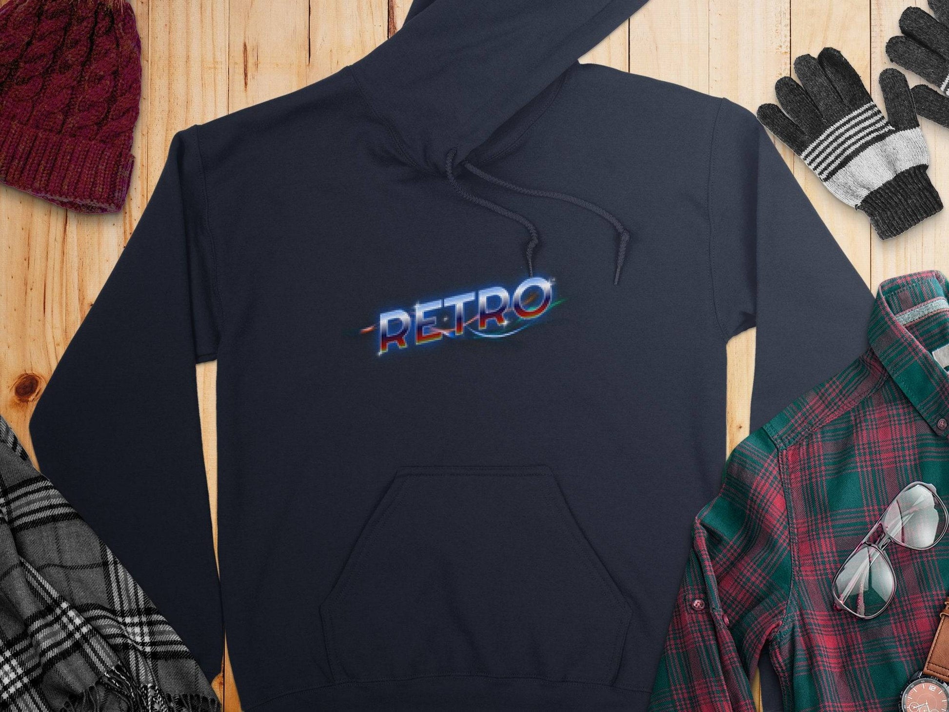 The Garment Graphics black hoodie with a vibrant neon Retro design is styled on a wooden surface, matched with a red beanie, black and gray gloves, a black and white plaid scarf, and a green and red plaid shirt for a modern trendy look.