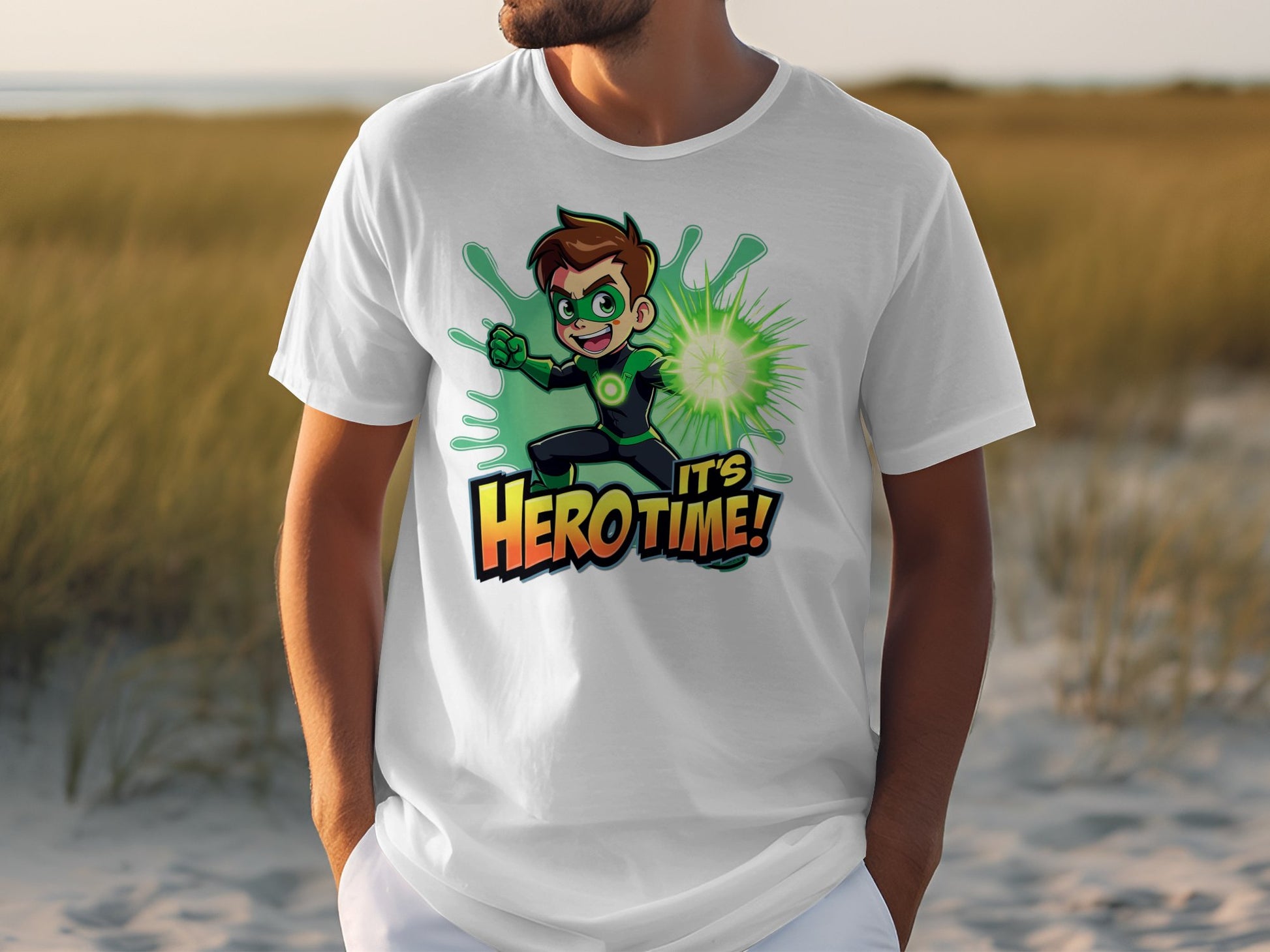 A person stands on sand, wearing a Garment Graphics vibrant white t-shirt that showcases a cartoon boy superhero with green energy and the phrase Its Hero Time! The superhero print vividly contrasts the blurred grass and sky background.
