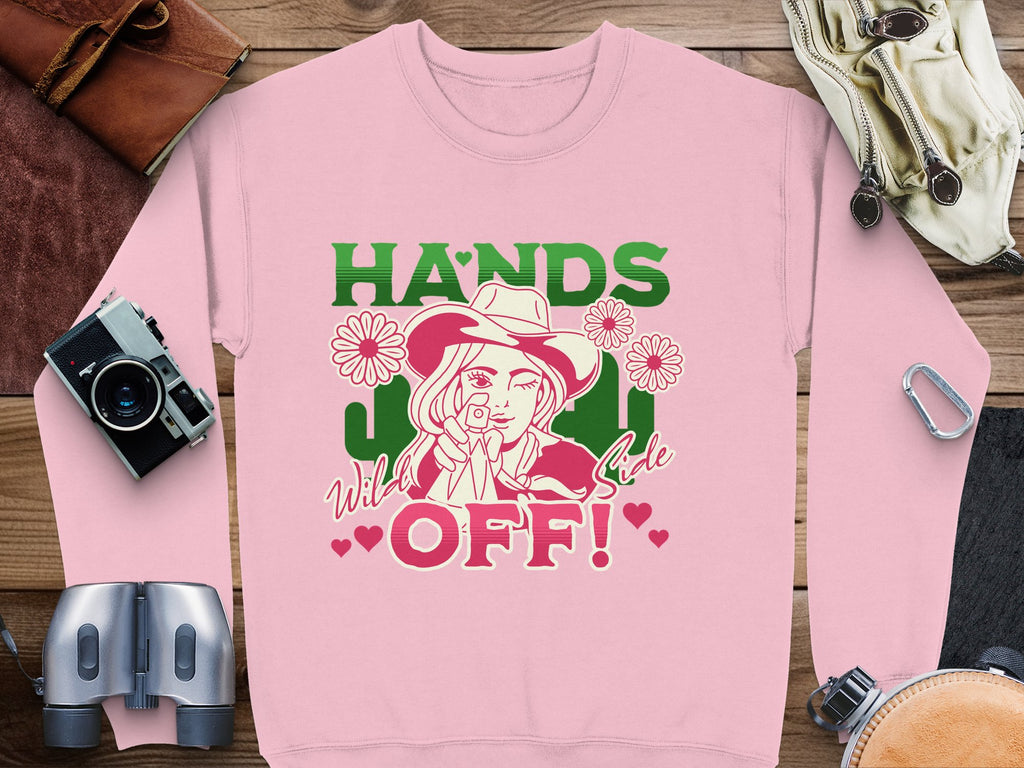 The Hands Off Wild Side Sweatshirt by Garment Graphics boasts a pink backdrop with striking green and red lettering, featuring a cowboy-hatted figure among flowers and hearts. Its loose fit is ideal for pairing with accessories like cameras, binoculars, and gloves for adventure seekers.