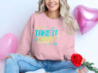 A person in a unisex Garment Graphics sweatshirt with the Take It Easy print and musical notes sits cross-legged, holding a red rose. Pink heart-shaped balloons float behind them, enhancing the relaxed vibe. Their ripped jeans highlight the casual style.