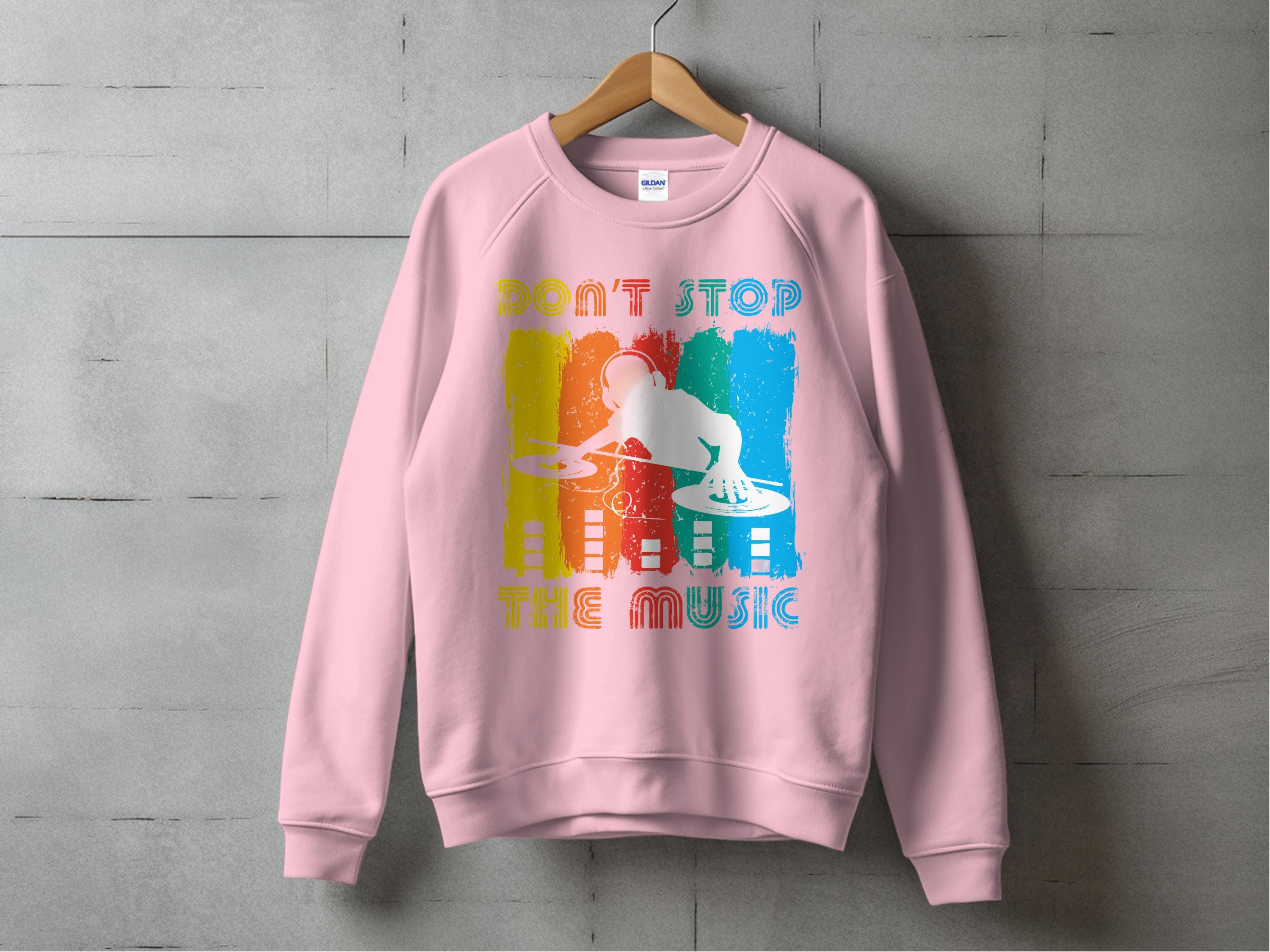 A pink Garment Graphics DJ sweatshirt, perfect for music lovers, hangs on a concrete wall. It features colorful graphics with a DJ silhouette at turntables and the phrase DONT STOP THE MUSIC, in blue, red, yellow, and white accents. Available in unisex sizing.