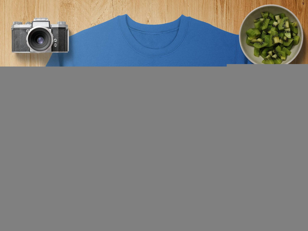 A blue sweatshirt by Garment Graphics features a green shamrock design with smaller shamrocks in a circle, capturing the Irish spirit. Its displayed on wood with a camera, lens, sliced kiwi bowl, burlap pouch, and map.