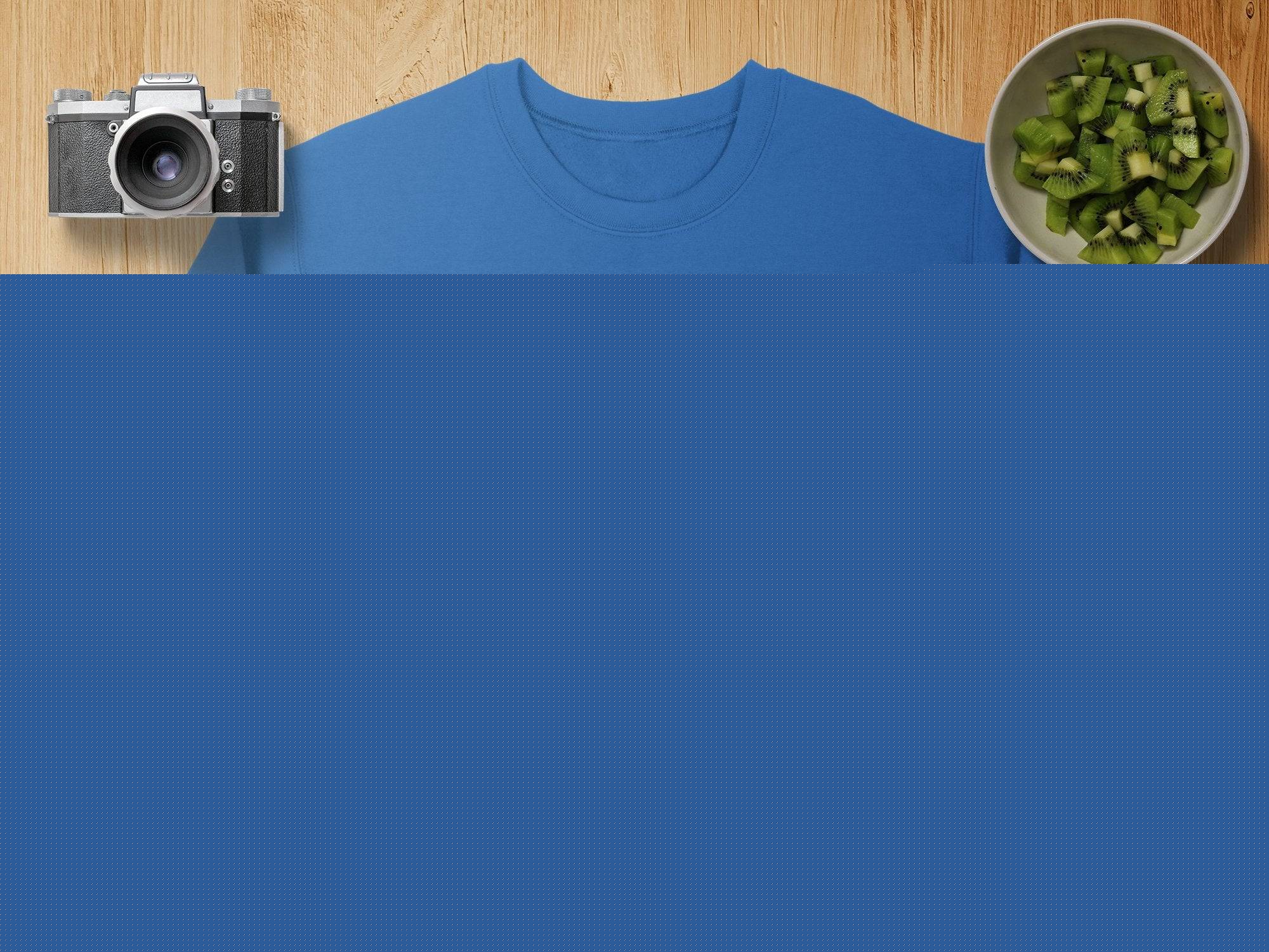 A blue sweatshirt by Garment Graphics features a green shamrock design with smaller shamrocks in a circle, capturing the Irish spirit. Its displayed on wood with a camera, lens, sliced kiwi bowl, burlap pouch, and map.